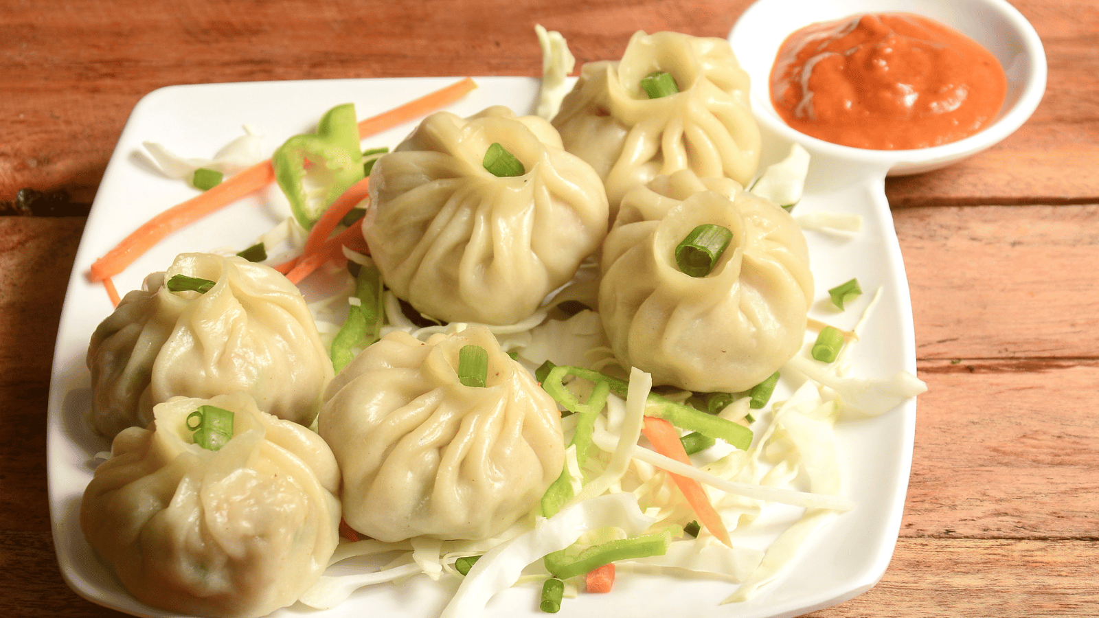 Six steamed dumplings on a bed of sliced carrots and cabbage sitting on white plate with red dipping sauce in corner