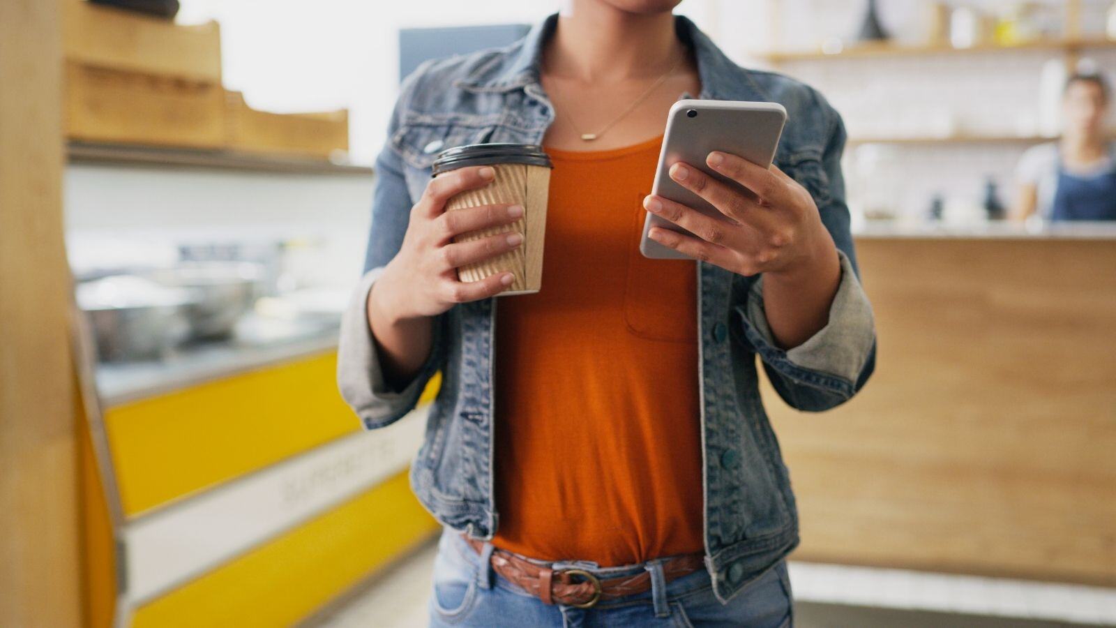 Woman in orange shirt and jean jacket holding phone in one hand and coffee cup in other hand. 